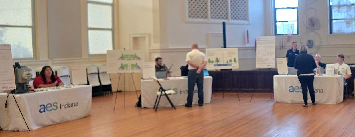 People interact at informational booths covered with AES Indiana tablecloths in a well-lit room with large windows and presentation boards. Some individuals are seated behind tables, while others stand and engage in conversation.