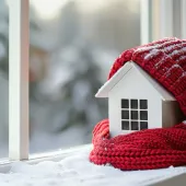Small White House Wrapped in a Red Knit Hat and Scarf Against a Snowy Window Background