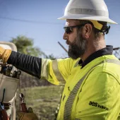 A utility worker wearing a yellow safety vest, gloves, a hard hat and protective glasses is reaching into a tool pouch. He is standing outdoors near a chain-link fence and utility lines, with a focused expression as he selects a tool. The worker's shirt features the "AES Indiana" logo, indicating his affiliation with the company. The background includes trees and clear skies