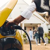  close-up of a yellow electric vehicle being plugged in to charge in a residential driveway. A person’s hand holds the charging connector, while in the background, a family, including two children, walks toward the entrance of a house. The scene highlights sustainable living with a focus on electric mobility and family life.