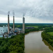 Aerial view of a power plant with three tall smokestacks, located near a river and surrounded by dense green forests and farmland. The facility includes large industrial buildings and equipment. The river winds through the landscape, and the cloudy sky adds a dramatic backdrop to the scene, highlighting industrial and natural elements.