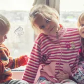 Three young children are sitting by a window, dressed in colorful winter sweaters. The child on the left, wearing an orange and blue sweater, is looking at the girl in the middle, who is wearing a pink striped sweater with a flower and soccer ball design. The girl on the right, dressed in a festive patterned sweater, is smiling at the camera. Snowflake decorations are visible on the window behind them.