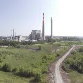 Wide aerial view of an industrial power plant with tall smokestacks, surrounded by green fields and a winding gravel road in the foreground under a clear sky.
