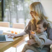 An adult seated at a table using a laptop while holding a small child on their lap. The child is holding a piece of fruit. A large window is in the background.