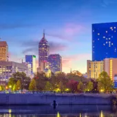 Indianapolis city skyline at dusk with illuminated buildings, the blue glass JW Marriott hotel, and reflections on the river in the foreground.