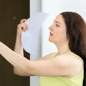A woman adjusting a thermostat on a wall while holding a piece of paper, appearing focused.