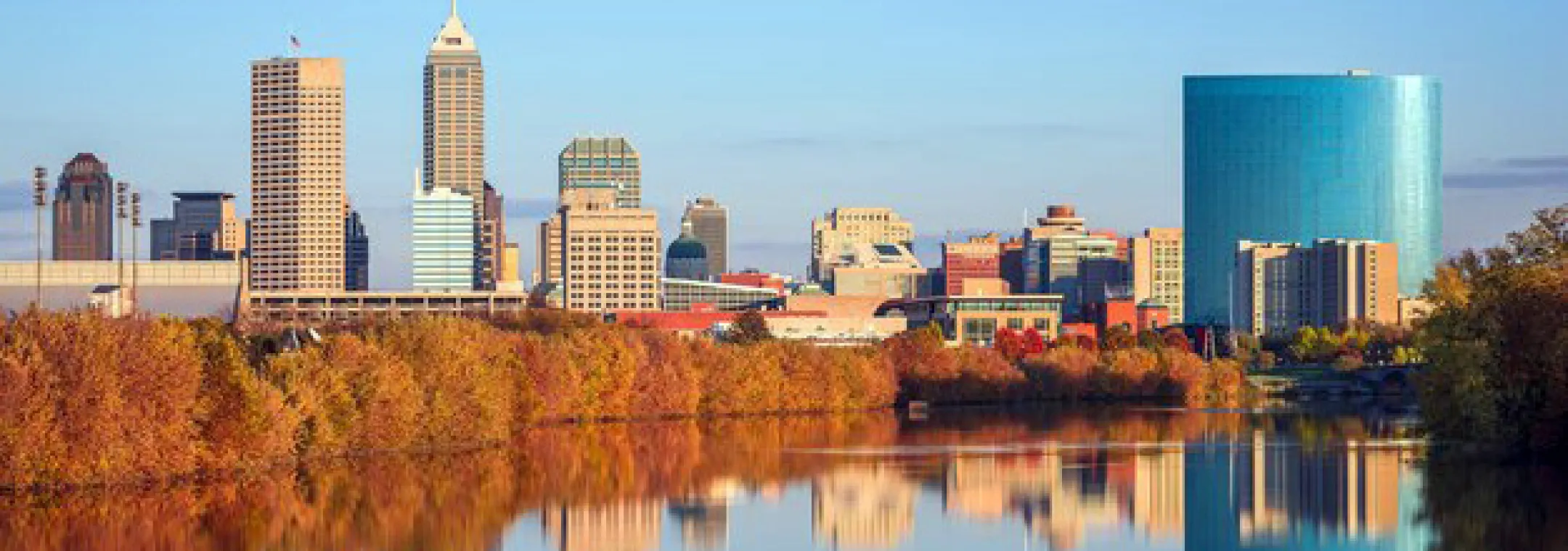 Downtown Indianapolis skyline with modern skyscrapers and a round blue building, reflected in a calm river bordered by autumn trees under a clear blue sky.