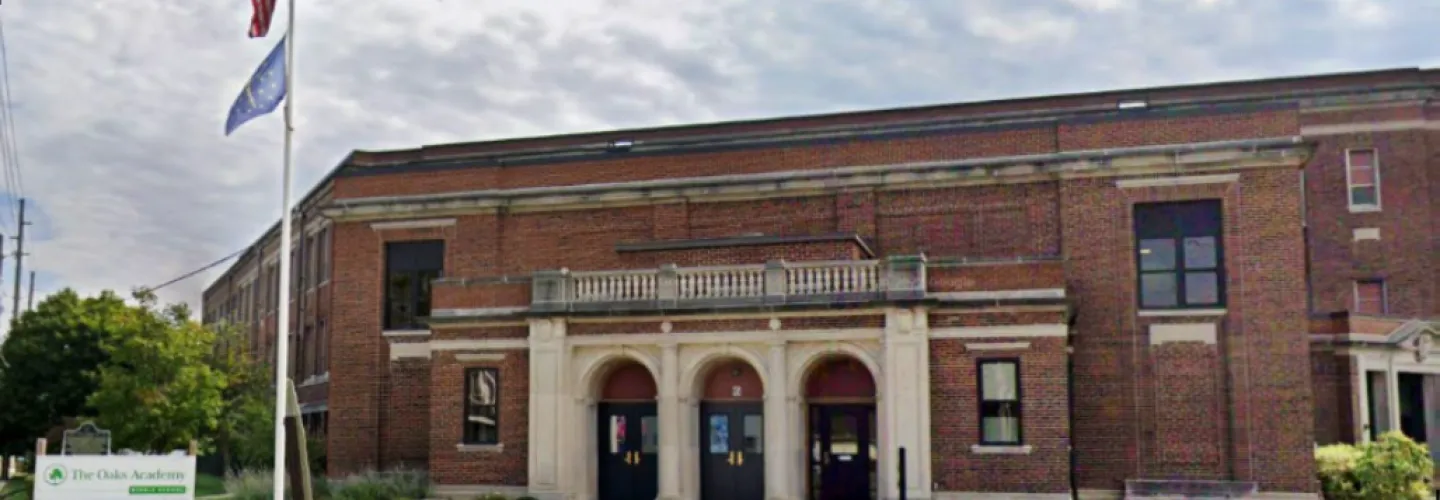 Front view of The Oaks Academy building in Indianapolis, Indiana, showing a historic brick school facade with entrance arches, flags, and educational signage under a partly cloudy sky.