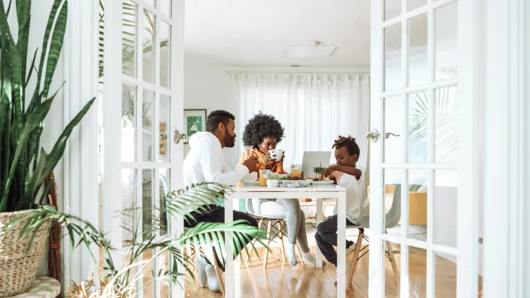 Two adults and one child seated at a dining table in a bright room, eating and using a laptop. Indoor plants and glass double doors frame the scene.