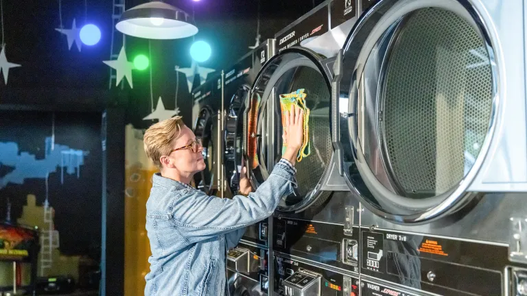 Person loading or unloading a large commercial dryer in a laundry facility, with multiple stacked machines visible and hanging decorations in the background.
