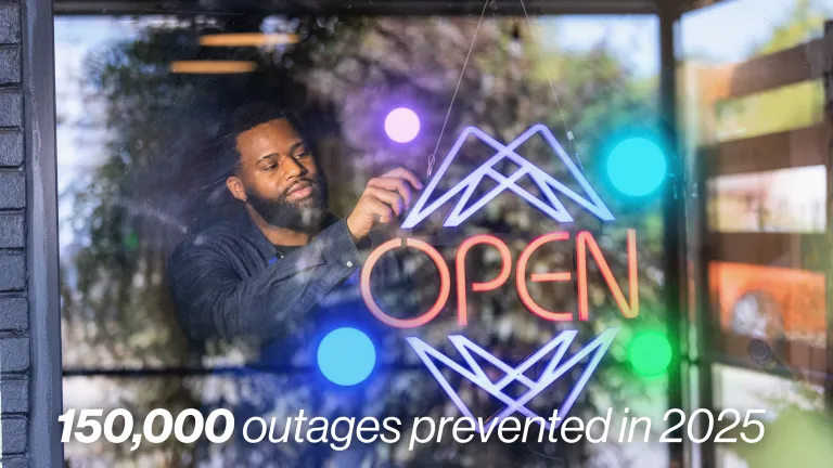 A man adjusts a glowing neon 'OPEN' sign in a storefront window with text reading '150,000 outages prevented in 2025'.