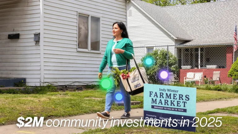 A woman carrying a tote bag of fresh vegetables walks past an Indy Winter Farmers Market sign near a residential home.