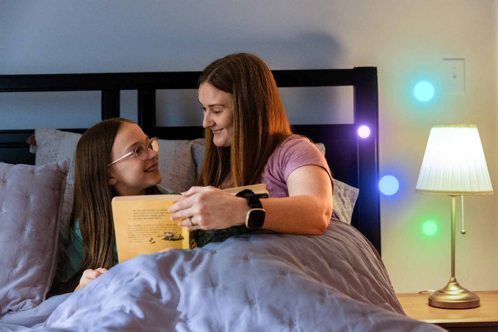 A mother and daughter smile while reading a book together in bed, with a lamp glowing softly beside them.