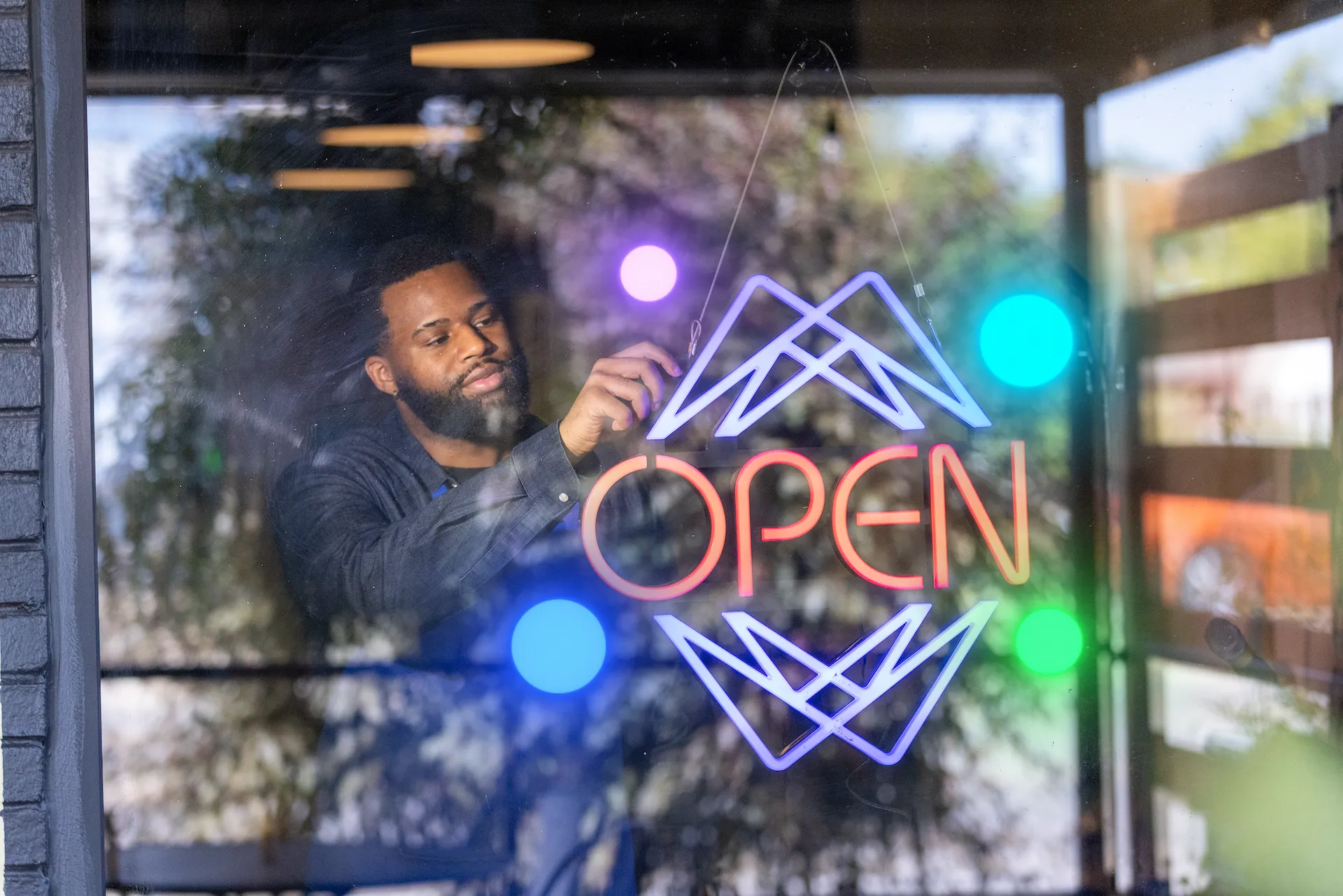 Person adjusting a neon ‘OPEN’ sign on a glass door with geometric designs, colorful glowing circles reflected on the glass.