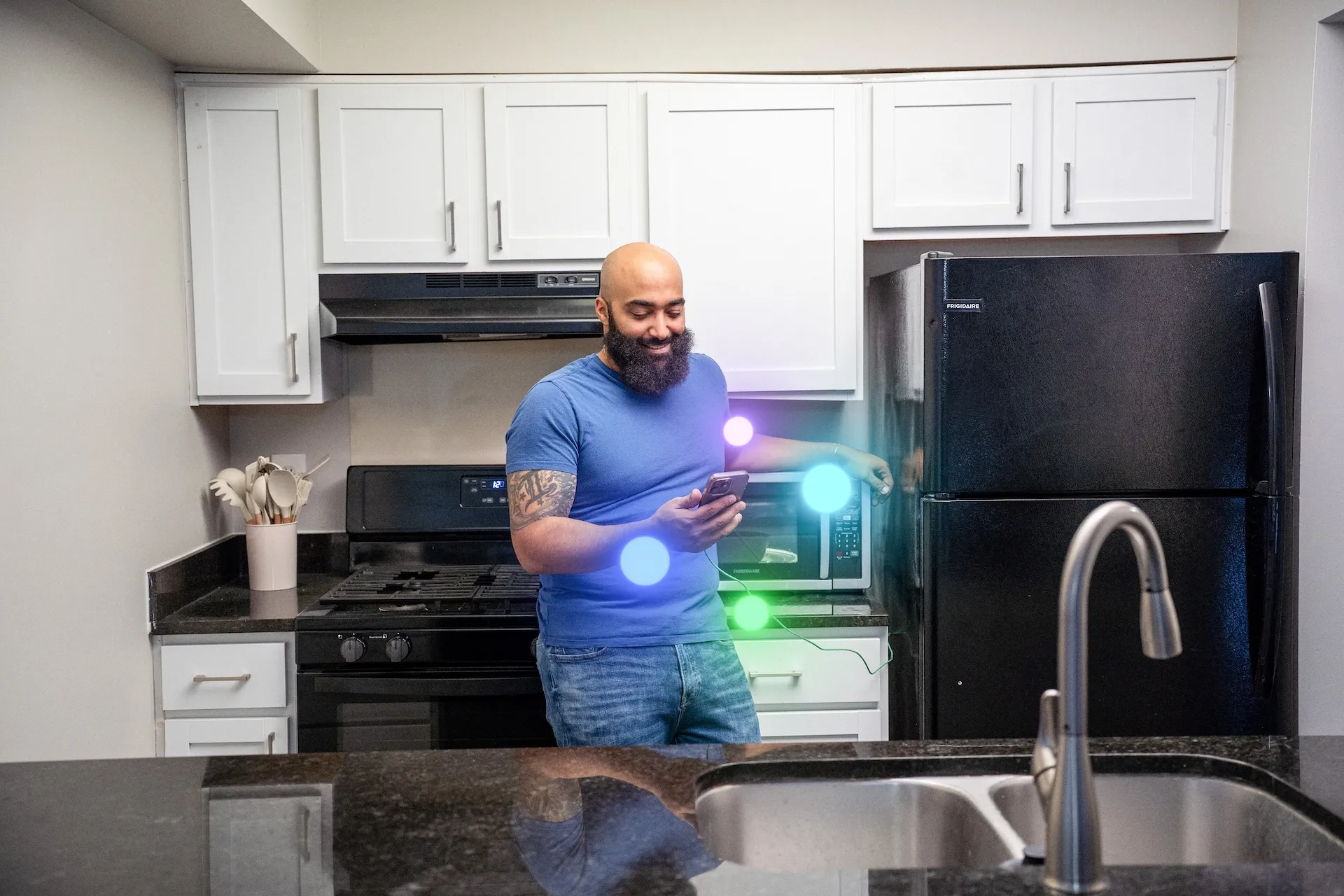 Person standing in a modern kitchen with white cabinets and black appliances, looking at a smartphone. Colorful glowing circles appear near the countertop and appliances.