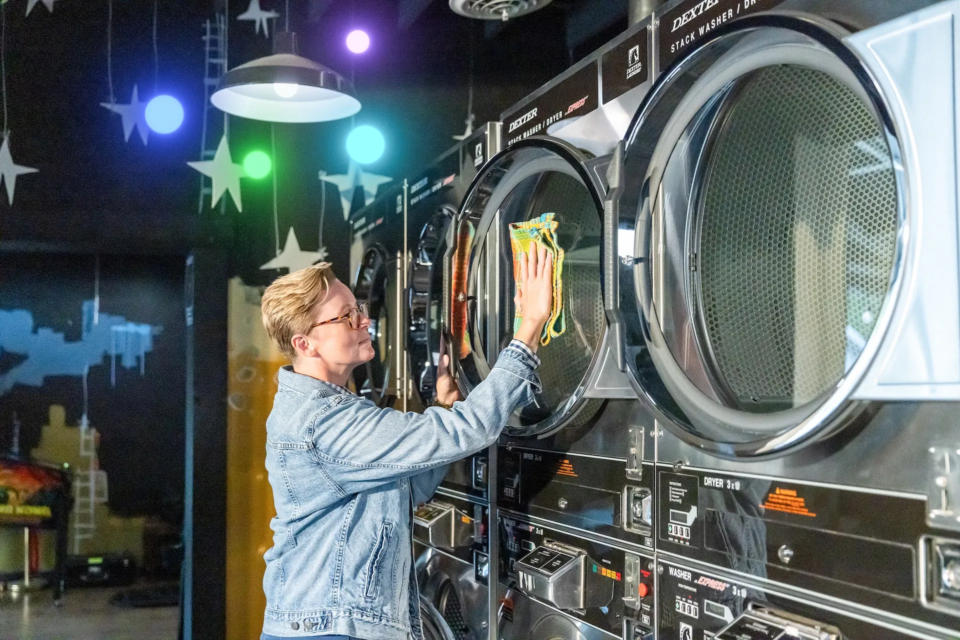 Person wearing a denim jacket cleaning the glass door of a large industrial dryer in a laundromat. The background features dark walls with star designs. Colorful glowing circles surround a hanging light.