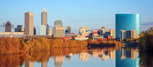 Downtown Indianapolis skyline with modern skyscrapers and a round blue building, reflected in a calm river bordered by autumn trees under a clear blue sky.