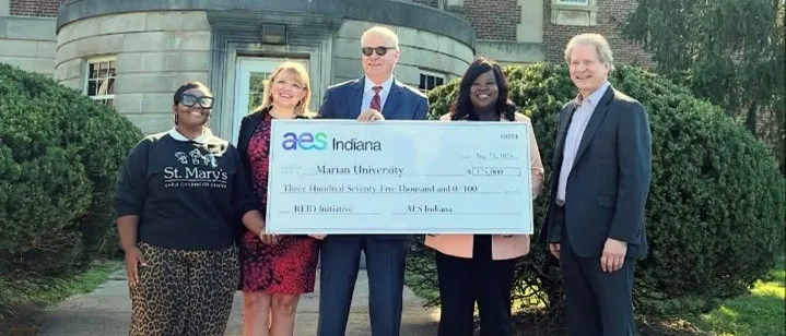 Five people stand outdoors in front of a building, smiling and holding a large ceremonial check from AES Indiana to Marian University for $375,000, supporting the READ Initiative.