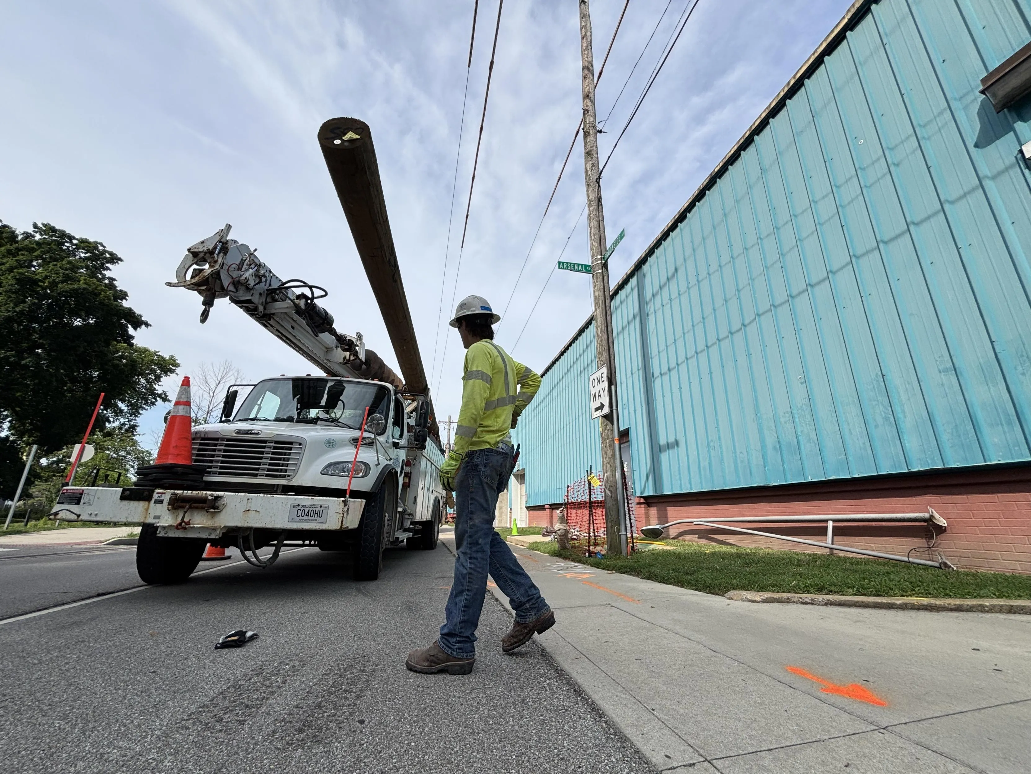A construction worker in a yellow safety vest and helmet stands near a utility truck lifting a wooden pole on a street. The truck is positioned next to a blue building with a one-way street sign visible.