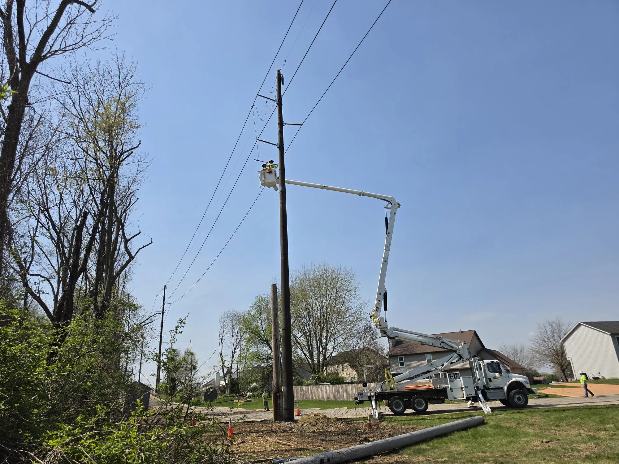 A utility worker in a bucket truck repairs power lines on a sunny day. The truck is parked on a residential street with houses and trees in the background.