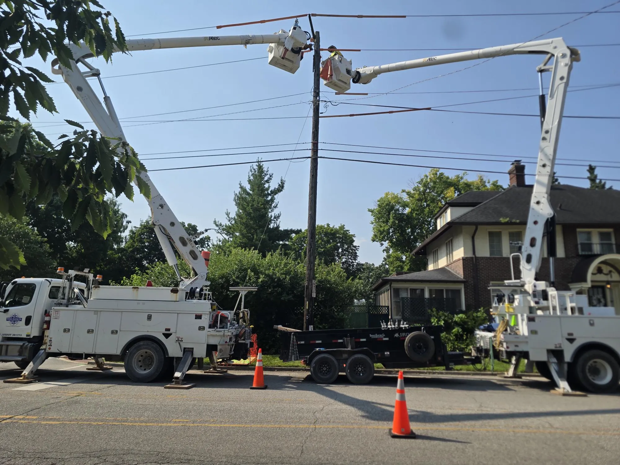 Two utility trucks with extended boom lifts on a residential street. Workers in safety gear are repairing power lines. Orange cones are placed around the trucks for safety.