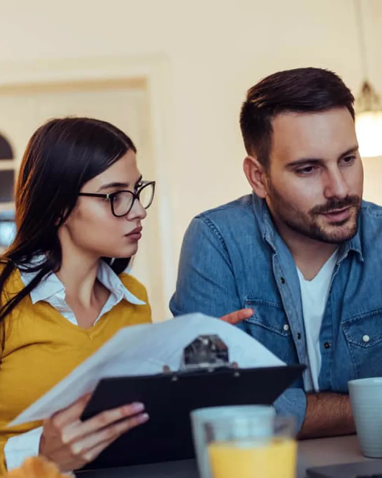 A young couple sits at a table, reviewing bills and financial documents. The woman, wearing glasses and a yellow sweater, holds a clipboard with papers, while the man, wearing a denim shirt, looks at a laptop screen with a focused expression. Both have coffee cups on the table, suggesting a discussion about household finances. The setting is a warmly lit home interior.