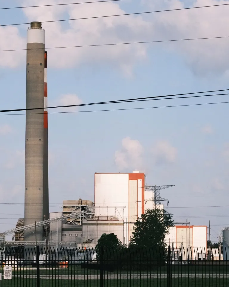 Industrial power plant facility with tall smokestacks, white and orange buildings, transmission towers, and security fencing under cloudy sky.