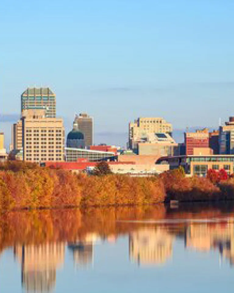 Downtown Indianapolis skyline with modern skyscrapers and a round blue building, reflected in a calm river bordered by autumn trees under a clear blue sky.