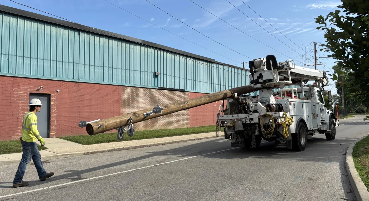 A utility worker in a yellow safety jacket walks beside a truck carrying a large wooden utility pole on a street. The truck is parked near a building with a red brick and teal metal exterior. Overhead power lines are visible against a blue sky.
