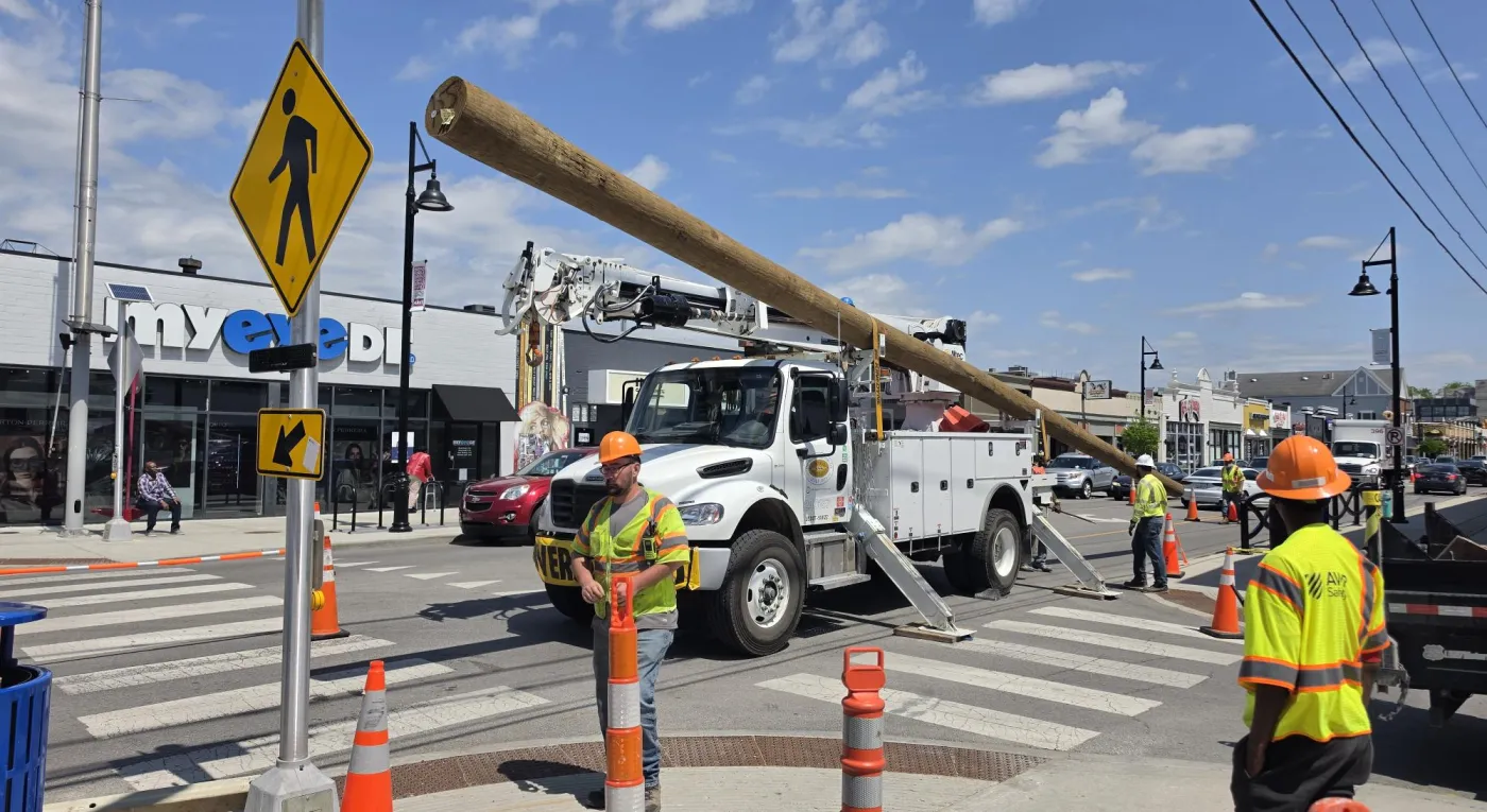 Construction workers in safety vests and helmets manage traffic around a utility truck lifting a wooden pole at a crosswalk in a busy street. Orange cones and a pedestrian sign are visible. Shops and pedestrians are in the background.