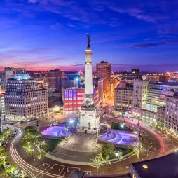 Aerial view of the Soldiers and Sailors Monument in downtown Indianapolis at night. The limestone monument stands at the center of a circular plaza, illuminated by bright lights. The surrounding buildings display colorful lighting, and streets curve around the monument with streaks of light from moving vehicles. The deep blue sky transitions into a warm sunset on the horizon, creating a vibrant cityscape.
