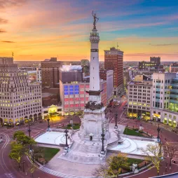 Aerial view of the Soldiers and Sailors Monument in downtown Indianapolis at sunset. The tall limestone monument, topped with a bronze statue, stands at the center of a circular plaza with fountains and sculptures. Surrounding the monument are historic and modern buildings, with city streets radiating outward. The sky displays warm hues of orange, pink, and blue, adding to the vibrant cityscape.
