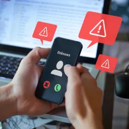 A person holding a smartphone displaying an incoming call from an unknown number, with red warning icons featuring exclamation marks appearing around the phone. A laptop screen with an open email inbox is visible in the background. The image suggests a potential phone scam or phishing attempt.