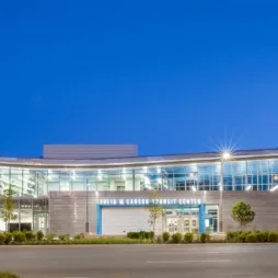 Evening view of the Julia M. Carson Transit Center, a modern glass and steel transportation hub with a curved roofline and bright exterior lighting. The center is surrounded by landscaped greenery and sits along a quiet street with the city skyline visible in the background. The sky is a rich blue, adding contrast to the illuminated building.