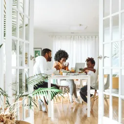 Two adults and one child seated at a dining table in a bright room, eating and using a laptop. Indoor plants and glass double doors frame the scene.