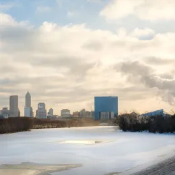 Winter scene of downtown Indianapolis, Indiana, showing the city skyline with Salesforce Tower and JW Marriott hotel. A frozen river with snow and ice covers the foreground. Industrial buildings on the right emit steam into a cloudy sky, highlighting the contrast between urban, natural, and industrial elements.