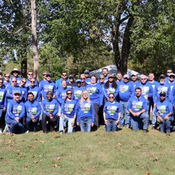 Large group of people wearing matching blue shirts with Day in the Parks printed on them, posing together outdoors in a park on a sunny day, with trees and event tents in the background.